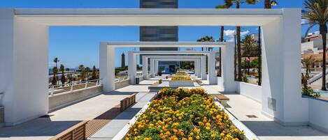 A modern architectural structure with a central garden of yellow flowers, white walls, and a clear view of the sea.