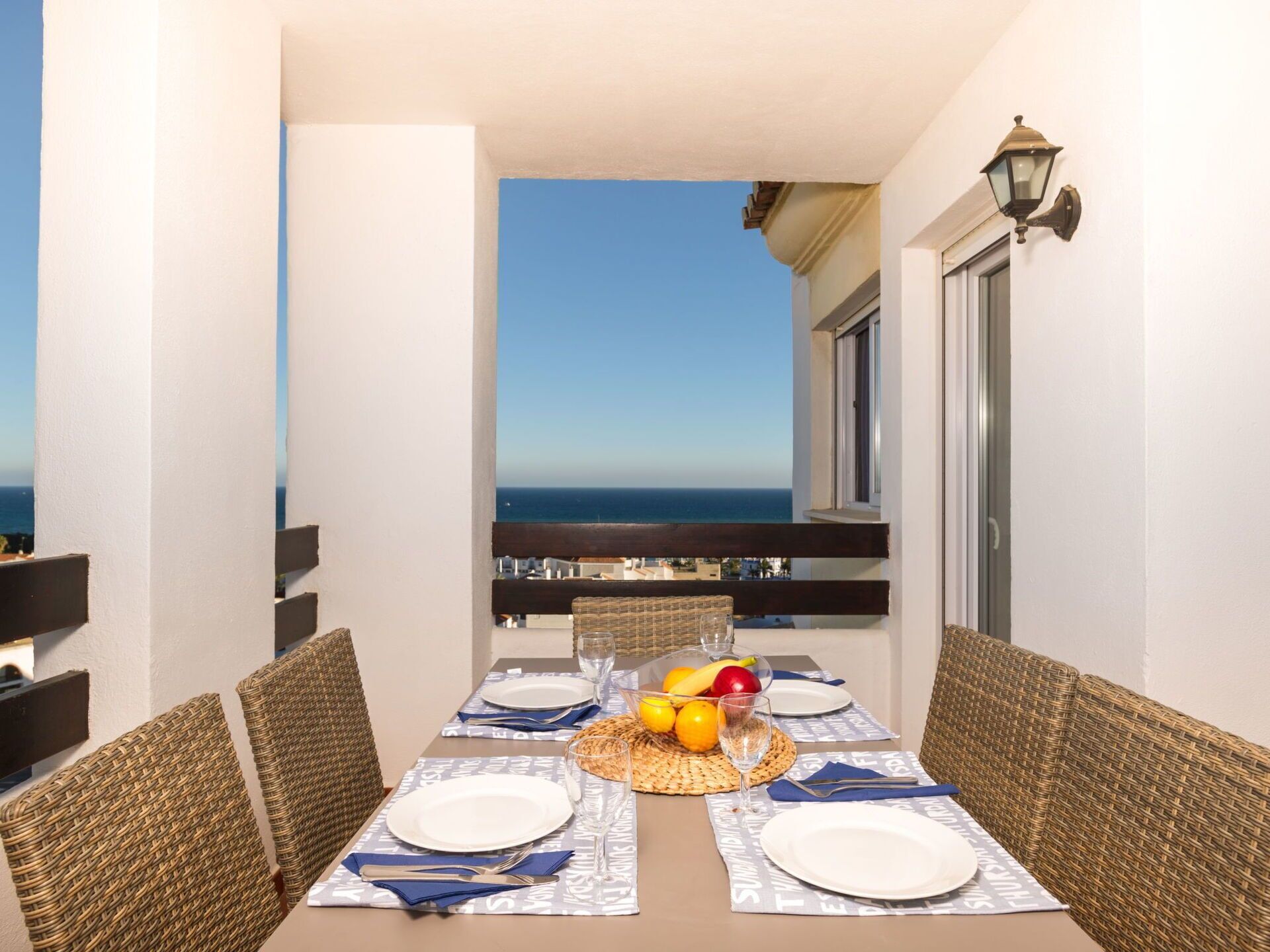 A dining table set for four with wicker chairs, a fruit bowl, and a view of the sea.