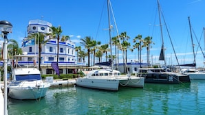 A marina with several yachts docked, a building with a blue and white facade, and palm trees in the background. - Nice apartment for 4 guests with A/C, pool, WIFI, TV and terrace (Estepona)