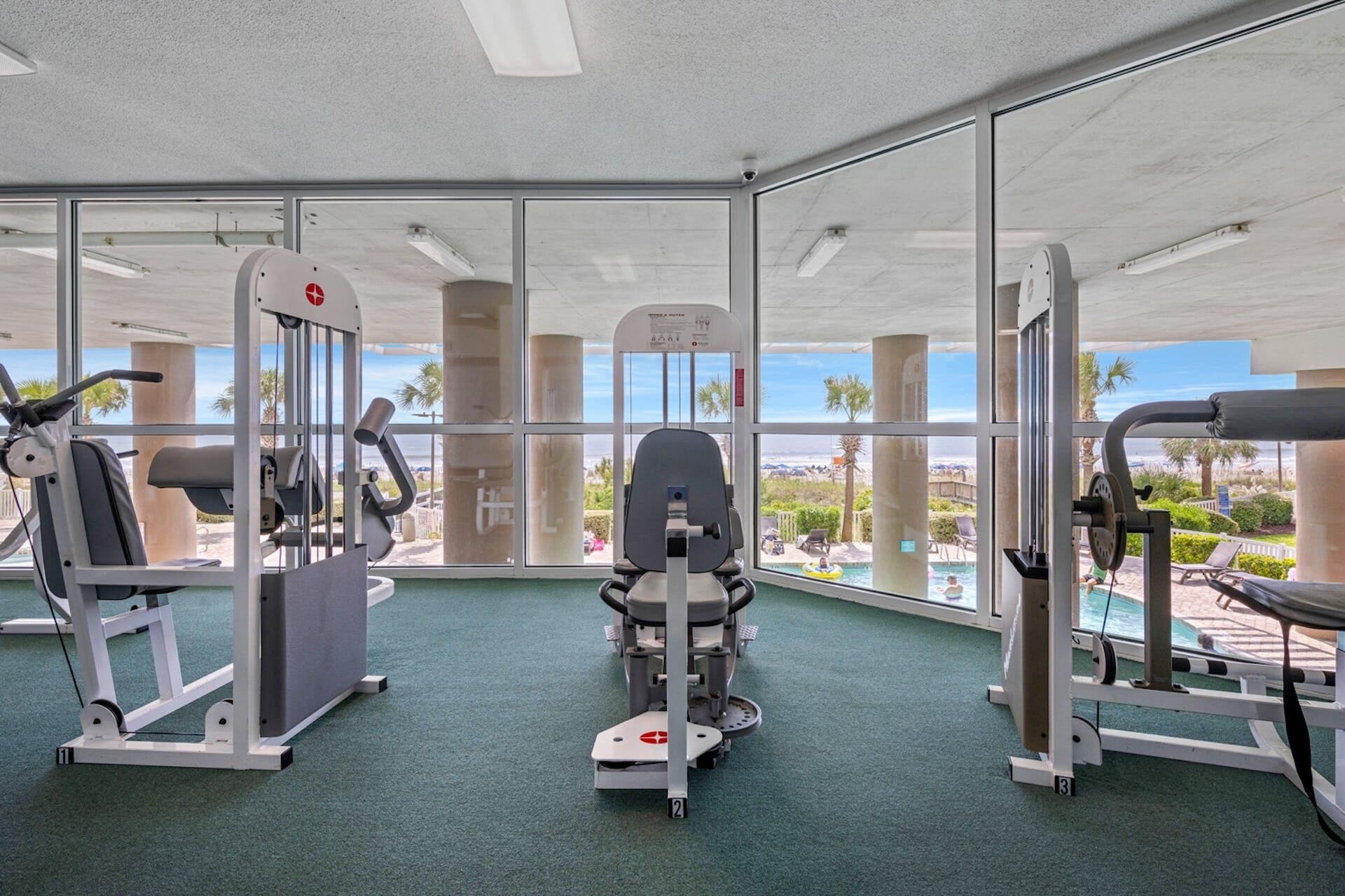 Ein Fitnessraum mit Trainingsgeräten und Blick auf ein Schwimmbad und Palmen.