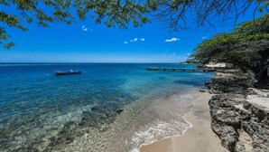 Une plage bordée d’une eau d’un bleu limpide, d’une côte rocheuse et d’une jetée qui s’avance dans la mer.