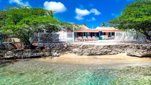 Une propriété située en bord de mer, avec une eau turquoise cristalline, une plage de sable et un bâtiment doté d’un toit rouge et de murs blancs.