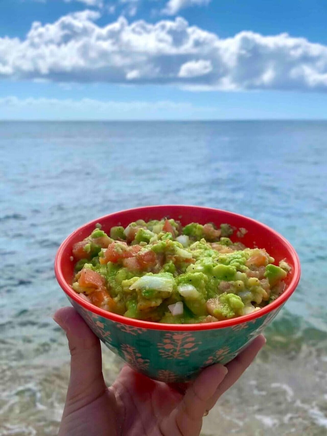 A bowl of guacamole held over a beach with clear water and a blue sky.