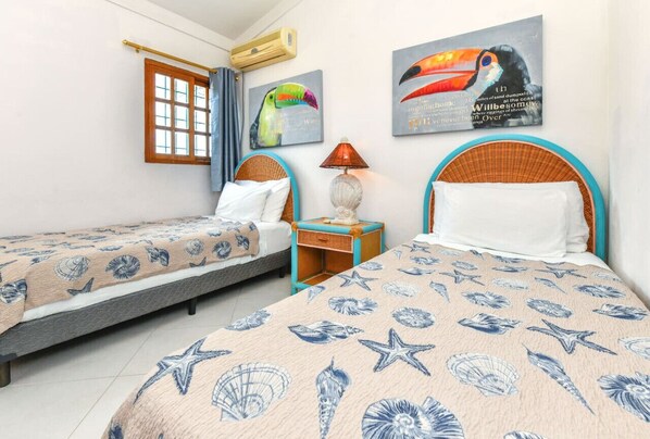 A hotel room with two beds, each with a headboard and a bedspread featuring sea creatures. There is a window with curtains, a wall-mounted air conditioner, and two wall art pieces depicting toucans.