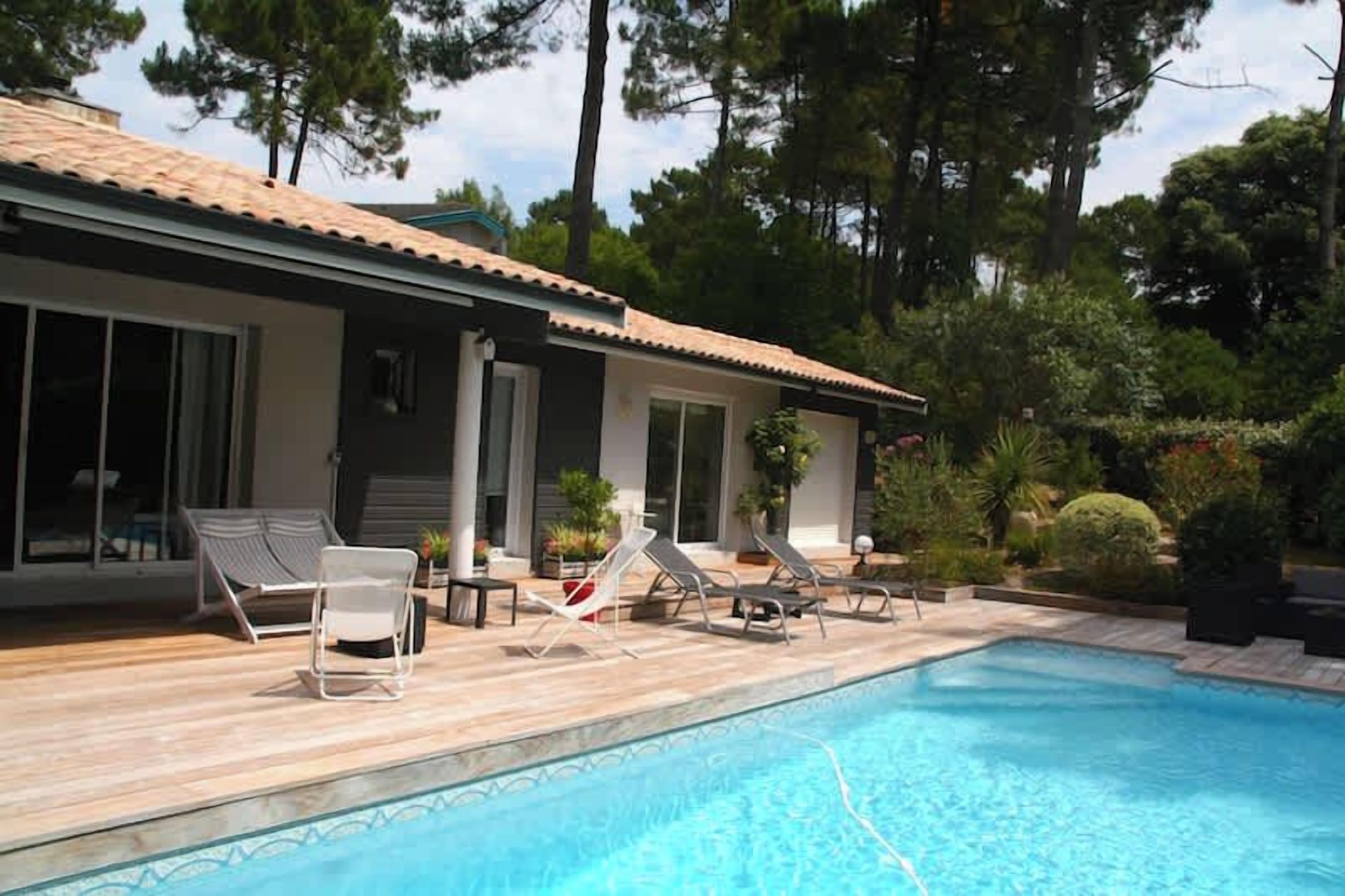 A swimming pool with lounge chairs, a covered deck, and a building with a tiled roof.