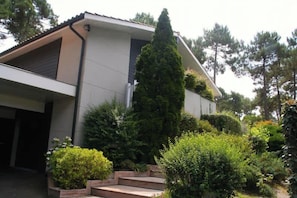 A modern two-story house with a sloped roof, surrounded by a well-maintained garden with various shrubs and a tall coniferous tree. - Comfortable house with pool G0269 (Lacanau)