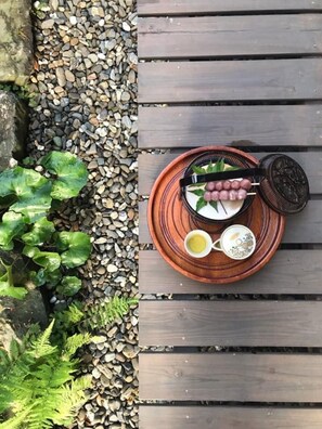 A wooden deck with a round table holding a plate with food and drinks, surrounded by pebbles and greenery.