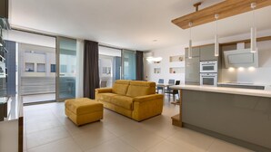 A modern living room with a sofa and ottoman, a kitchen area with a TV and oven, and large windows with curtains.