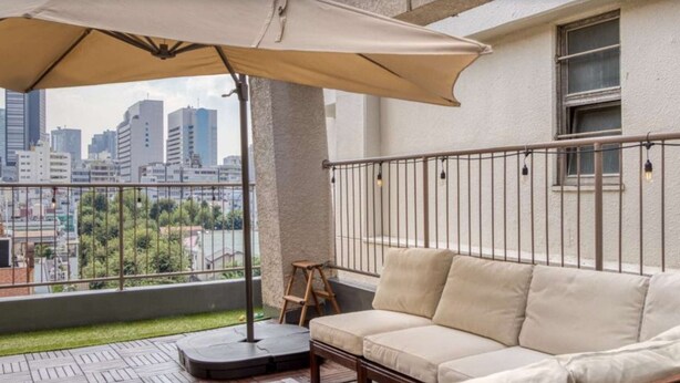 A balcony with a beige sofa, a small wooden stool, and a table. A large umbrella provides shade. The balcony overlooks a cityscape with tall buildings.