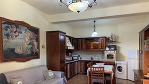 A living room with a sofa, a dining table with chairs, a kitchen with wooden cabinets, a washing machine, and a ceiling light fixture. - Apartment/ flat - SICULIANA House with garden (SICULIANA)