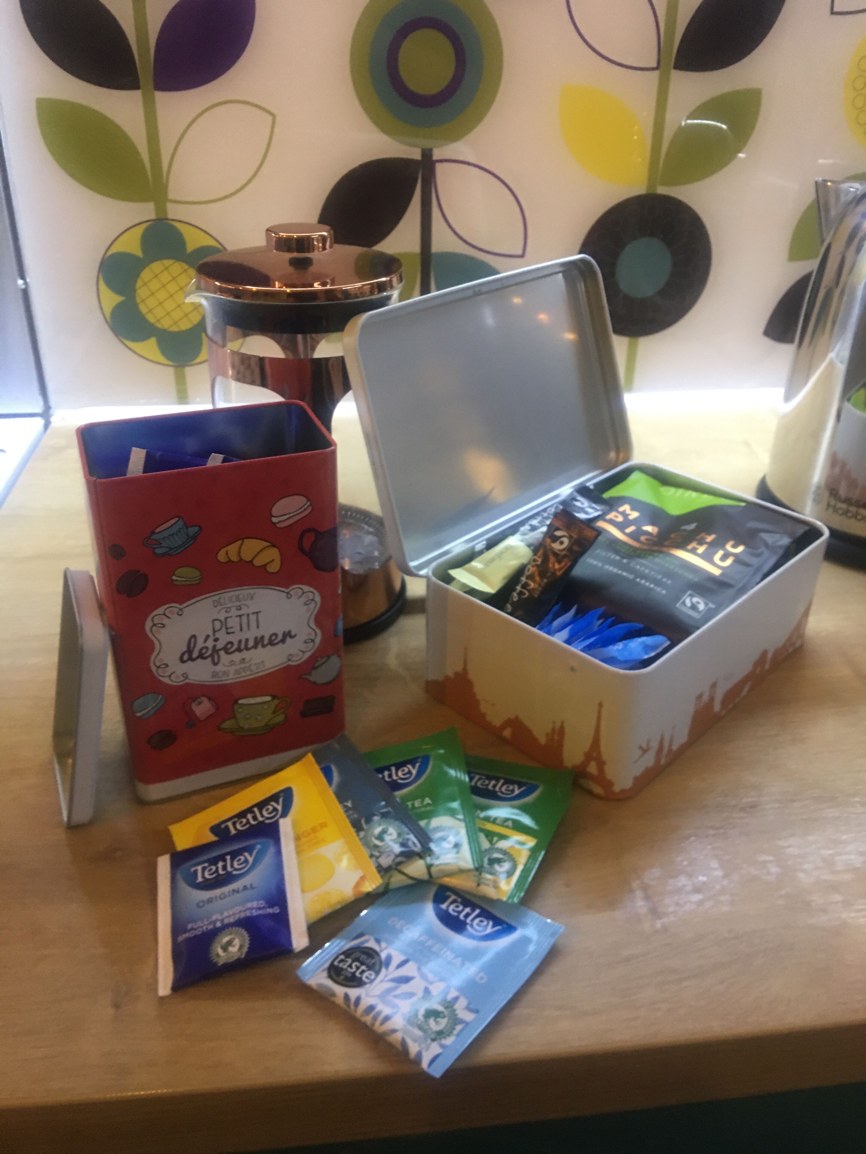 Une table avec un récipient rouge étiqueté « Petit déjeuner », une boîte de sachets de thé Tetley et une machine à café.