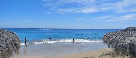 A beach with thatched umbrellas, clear blue water, and footprints in the sand.