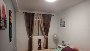 A bedroom with a bed, a bedside table with a lamp, a fan, and a window with curtains.