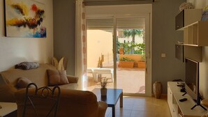 A living room with a sofa, a coffee table, and a TV stand. There is a sliding glass door leading to an outdoor area with plants and a table.