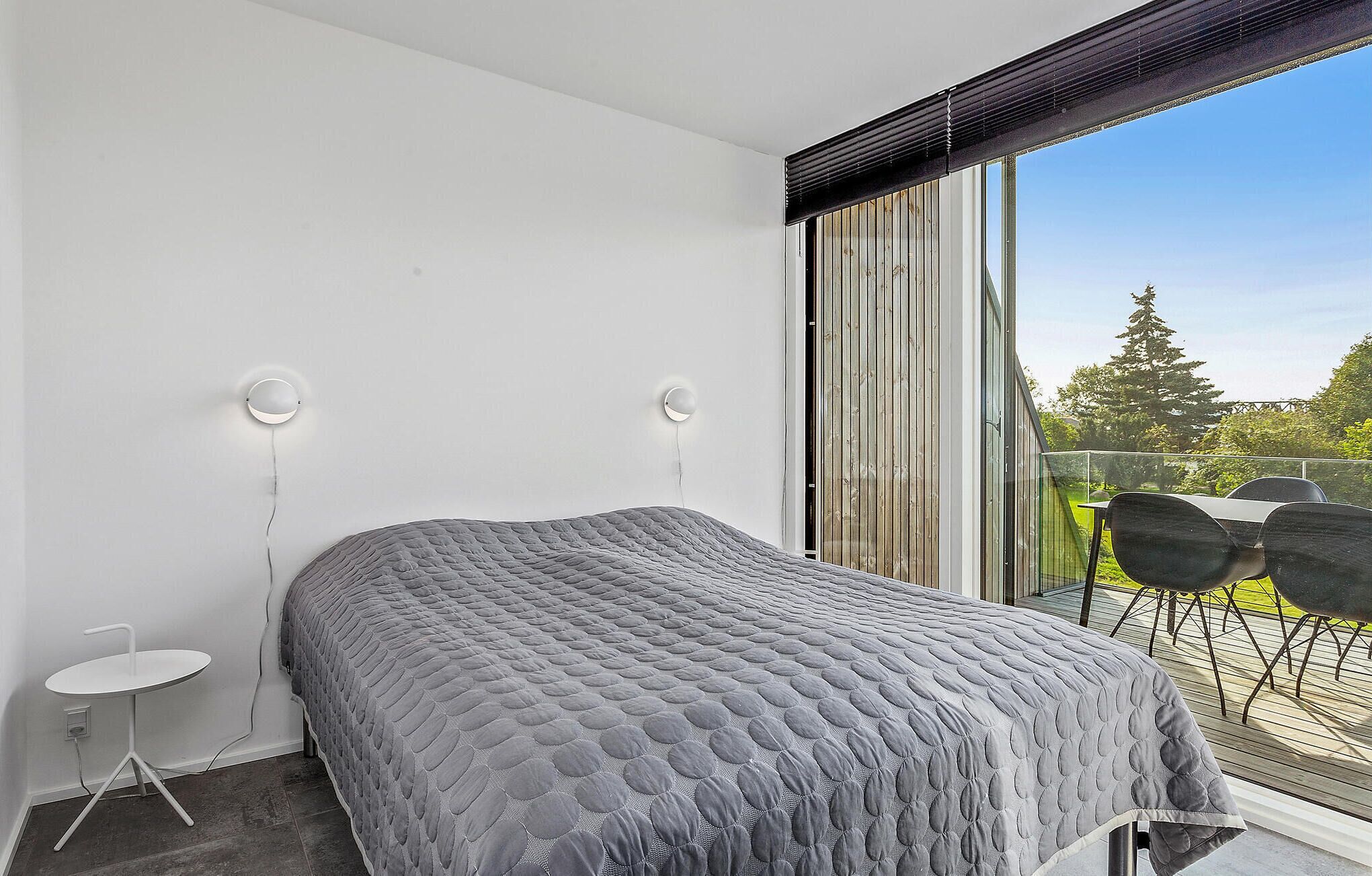 Ein Schlafzimmer mit einem großen Bett, zwei Wandleuchten, einem kleinen Tisch und Blick ins Freie durch eine Schiebetür.