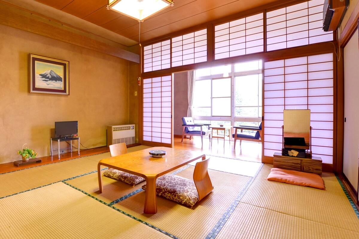 Una tradizionale stanza giapponese con pavimento in tatami, un tavolino, sedie, televisore e un'ampia finestra con paraventi shoji.