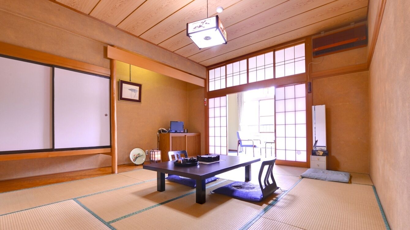 Um quarto japonês tradicional com tatames, uma mesa baixa com almofadas, uma escrivaninha com computador e portas deslizantes.