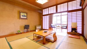Ein traditioneller japanischer Raum mit Tatami-Boden, einem niedrigen Tisch, Stühlen, einem Fernseher und einem großen Fenster mit Shoji-Schirmen.