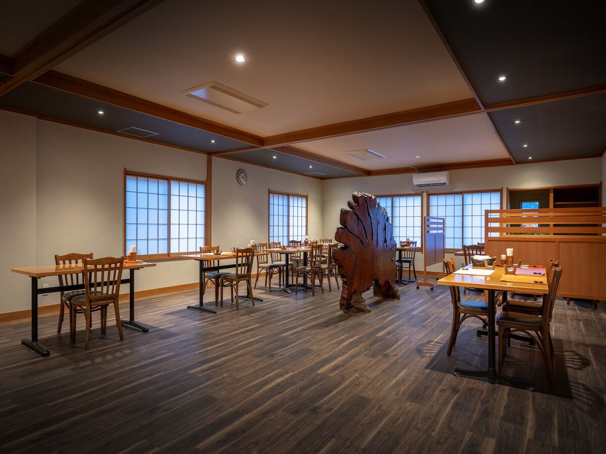 A spacious room with multiple wooden tables and chairs, large windows, and a central wooden sculpture.