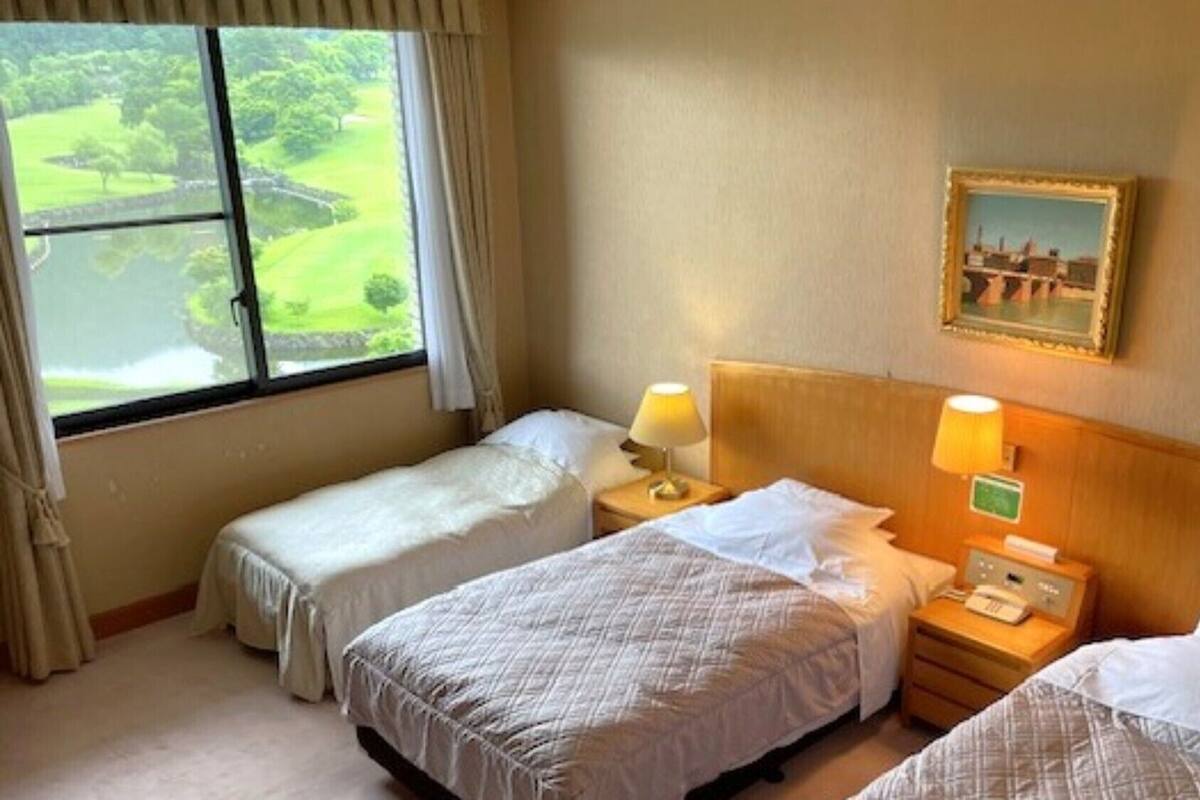 A hotel room with two beds, a window with a view of a golf course, a painting on the wall, and a bedside table with a lamp.