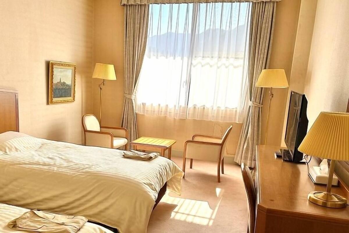 A hotel room with a bed, two chairs, a desk, a lamp, and a painting on the wall.