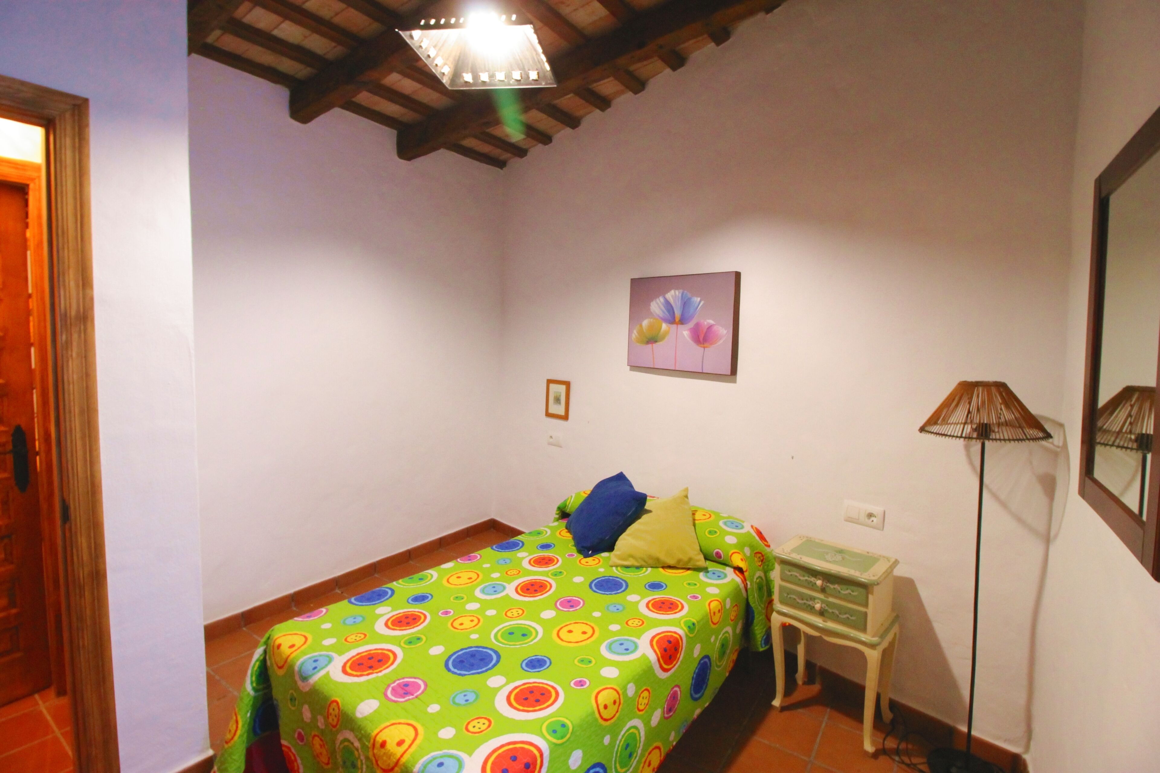 Una camera da letto con un letto ricoperto da una coperta dai colori vivaci e fantasia a cerchi, un comodino con una lampada e un quadro appeso al muro.