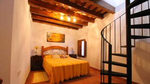 A bedroom with a wooden headboard, a bed with a yellow bedspread, a nightstand with a lamp, a painting on the wall, and a spiral staircase.