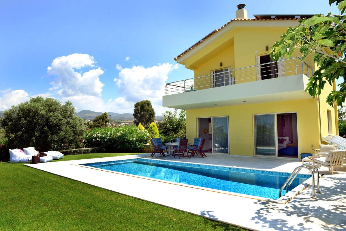 A two-story yellow house with a swimming pool, outdoor furniture, and a view of the landscape.