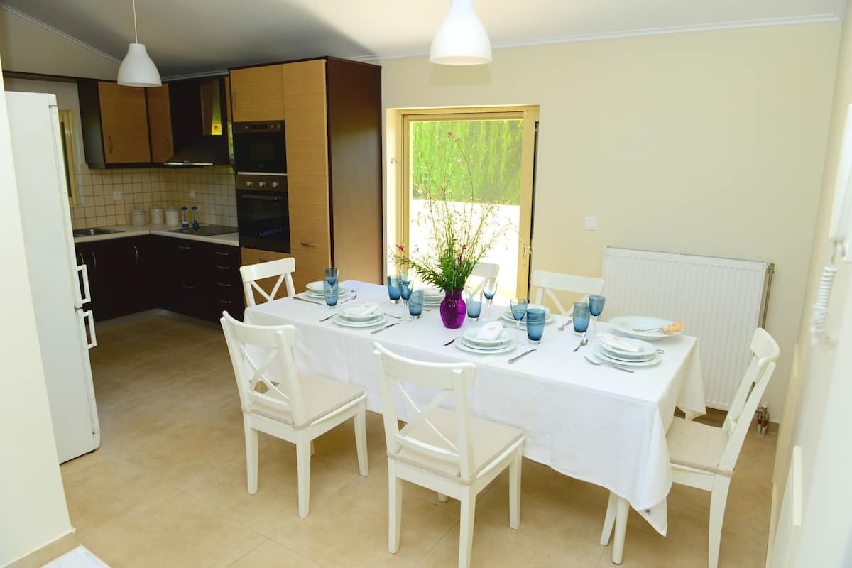 A dining area with a white table set for four, surrounded by white chairs. A vase with flowers is on the table, and there is a window with a view of greenery outside.