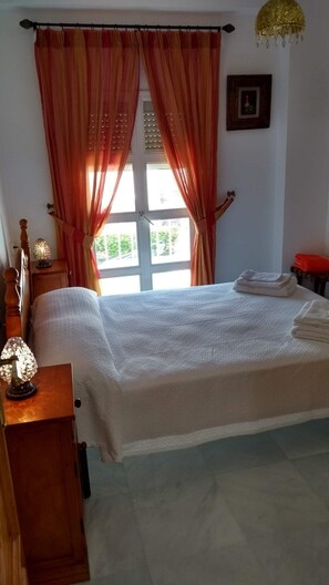 A neatly made bed with white linens, a wooden nightstand with a lamp, and a window with orange curtains. - NERJA SOLYMAR APARTMENTS (Nerja)