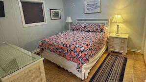 Une chambre à coucher comprenant un lit, un ventilateur de plafond, une table de chevet, une commode, une fenêtre avec des stores et un tapis.