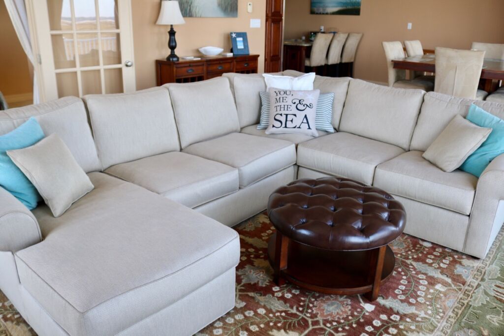 Un canapé d’angle beige avec un pouf capitonné, un tapis à motifs floraux et un coussin décoratif portant l’inscription « TOI, MOI, LA MER ».