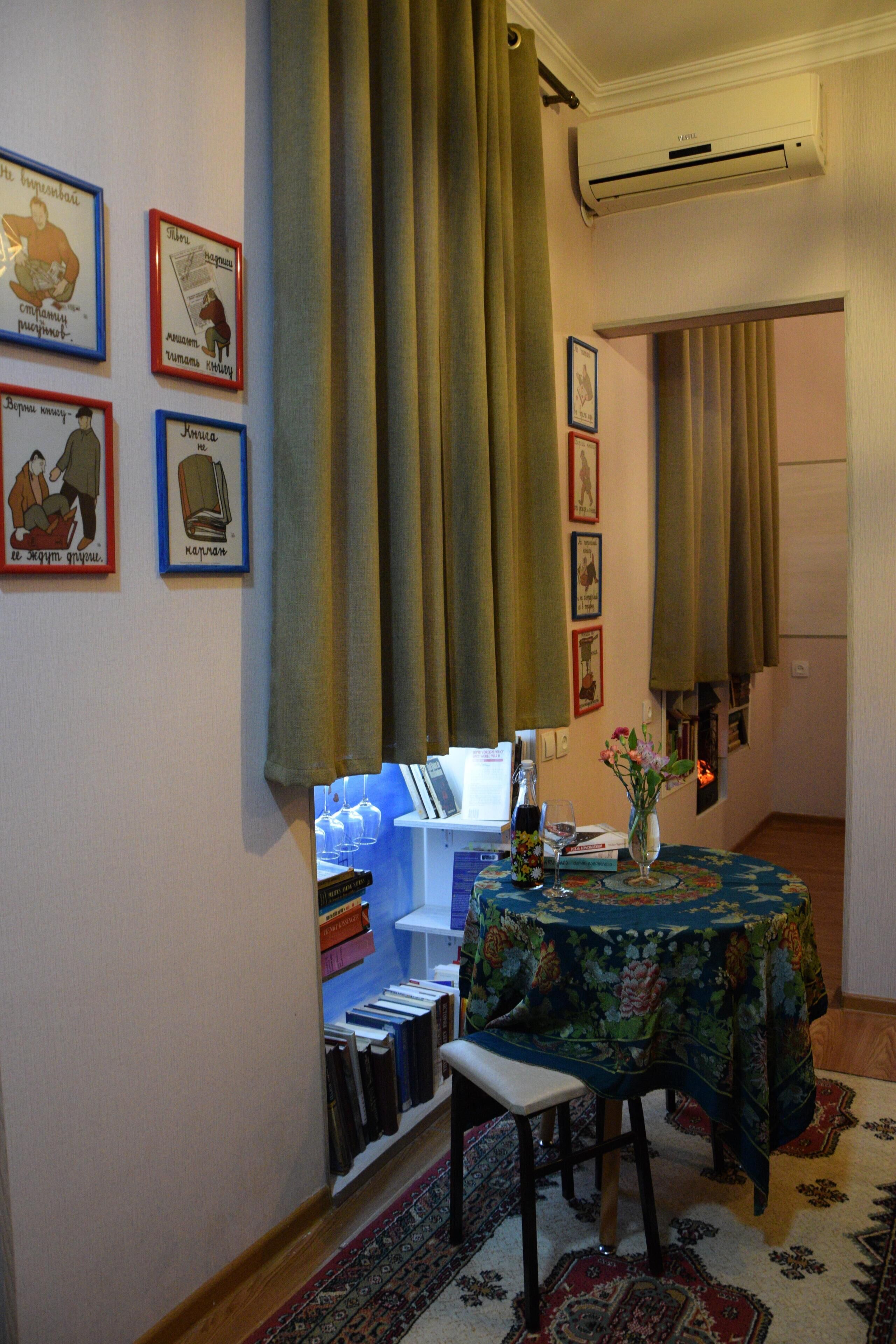 A room with a dining table covered with a floral tablecloth, a bookshelf, and a vase with flowers.