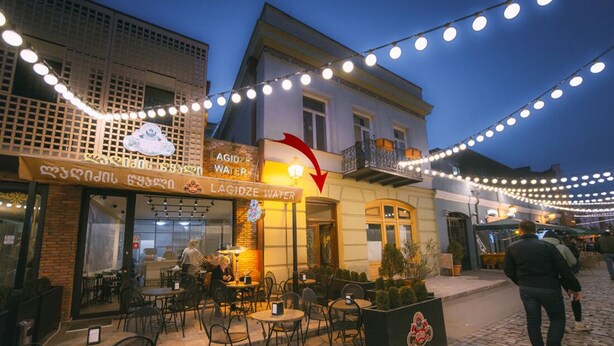 An outdoor café with tables and chairs, a sign for 'LAGIDZE WATER', and string lights. - The Librarians' Flat (Dzveli Tbilisi)