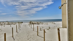 Een zandpad aan het strand, met houten palen en touwen, dat leidt naar een gebouw met een zichtbare douchezone.