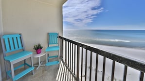 Een balkon met twee turquoise stoelen en een kleine tafel, uitkijkend op een strand met een heldere lucht.