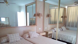 Une chambre d’hôtel avec deux lits simples, un grand miroir, un ventilateur de plafond et une fenêtre avec des rideaux.