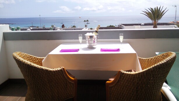 Une table avec une bouteille de champagne, deux verres et une serviette violette, sur un balcon donnant sur la mer.