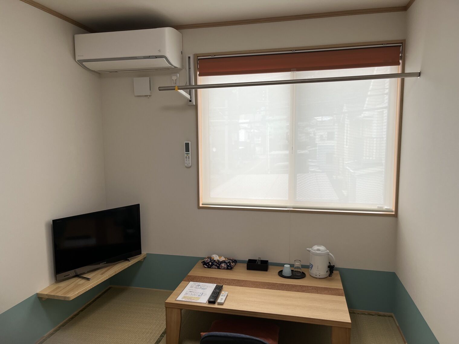 Una habitación pequeña con una televisión en un estante, una mesa con un hervidor y controles remotos, una ventana con persianas y un aire acondicionado.
