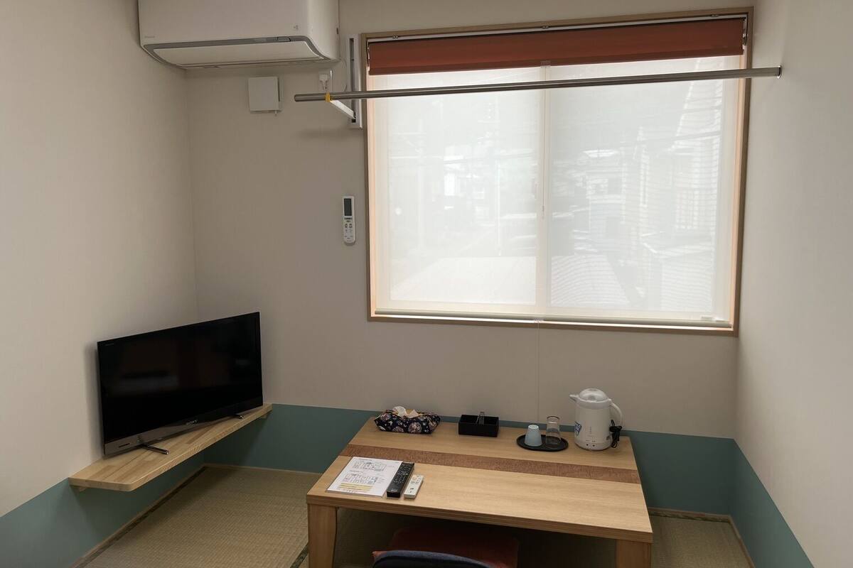 Una habitación pequeña con una televisión en un estante, una mesa con un hervidor y controles remotos, una ventana con persianas y un aire acondicionado.