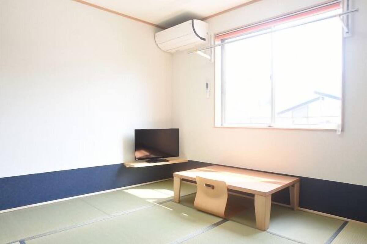 Una habitación con suelo de tatami, un televisor sobre un mueble bajo, una ventana con vistas y un aire acondicionado instalado en el techo.