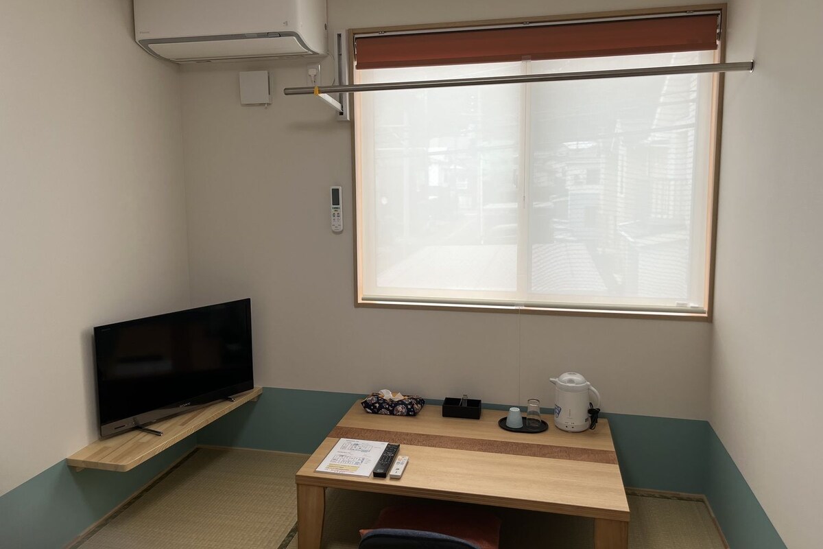 A small room with a TV on a shelf, a table with a kettle and remote controls, a window with blinds, and an air conditioner.