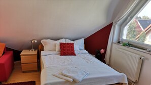 Une chambre avec un lit en bois, une literie blanche, un coussin rouge, une lampe de chevet, une chaise rouge et une fenêtre donnant sur des immeubles.