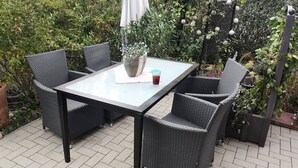 Une terrasse dotée d’une table à plateau en verre et de chaises en osier, entourée de verdure et d’une clôture décorative.