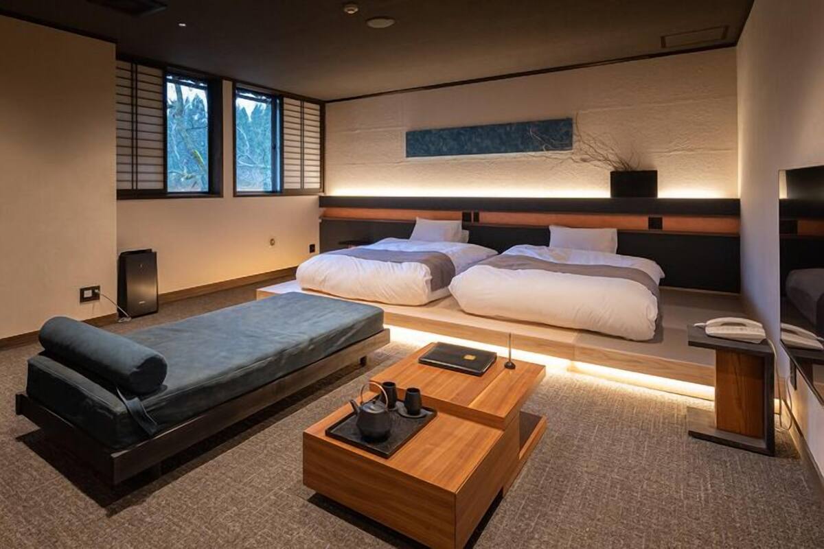 A modern hotel room with a sofa, two beds, a wooden coffee table, and a large window with a view.