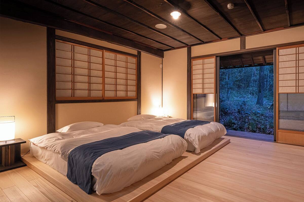 A traditional Japanese-style room with tatami flooring, a low wooden bed, sliding shoji doors, and a view of a forest.