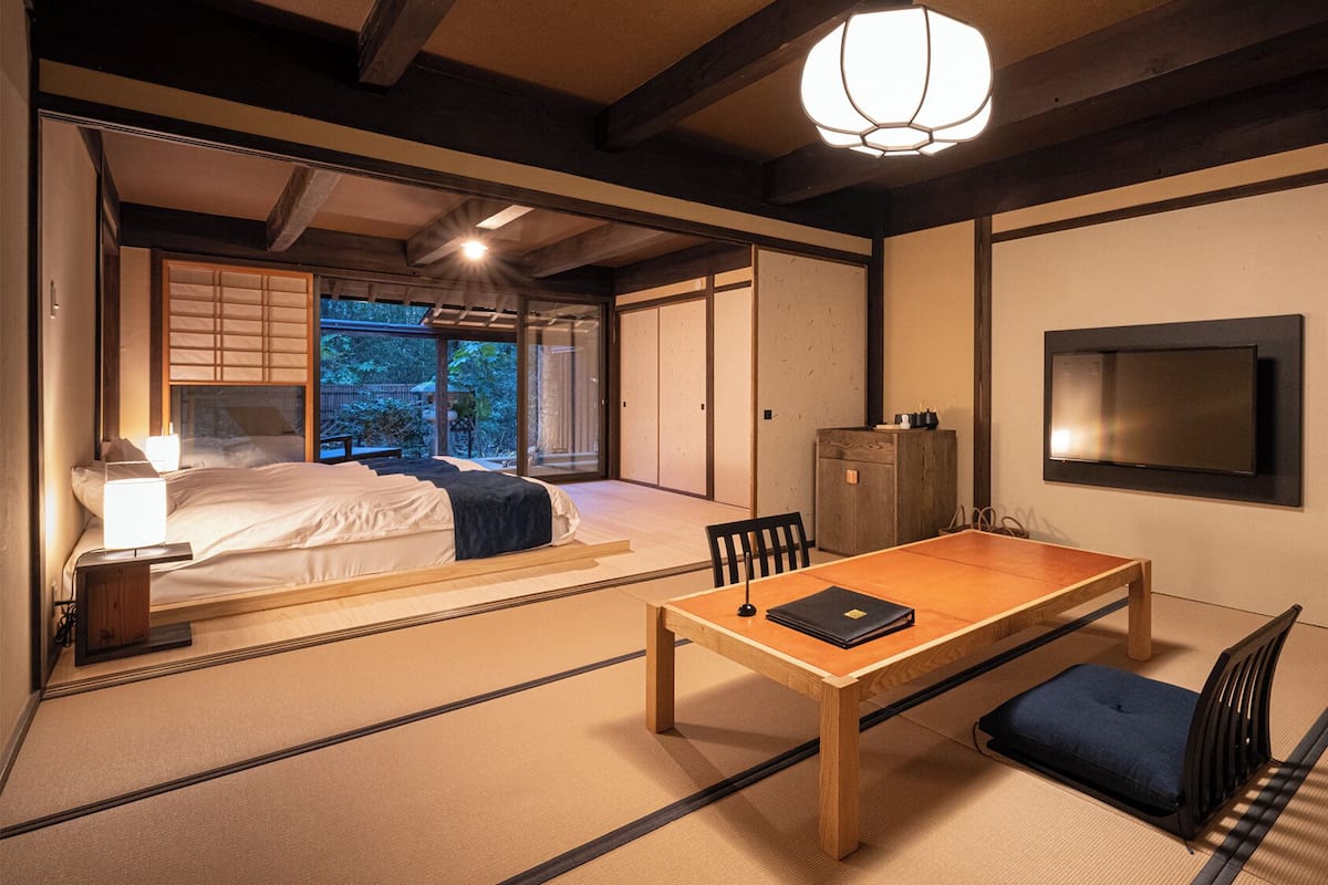 A traditional Japanese-style room with tatami flooring, a low table, a bed with a view to the outside, and a television mounted on the wall.