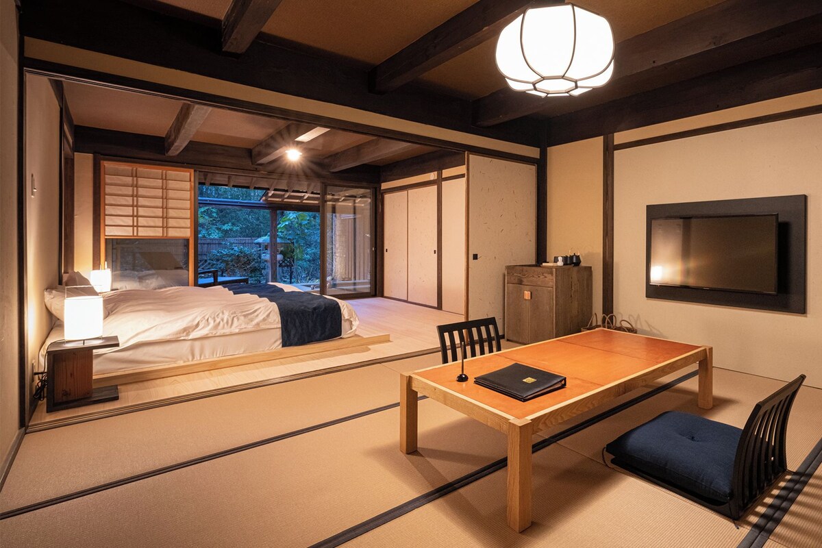 A traditional Japanese-style room with tatami flooring, a low table, a bed with a view to the outside, and a television mounted on the wall.