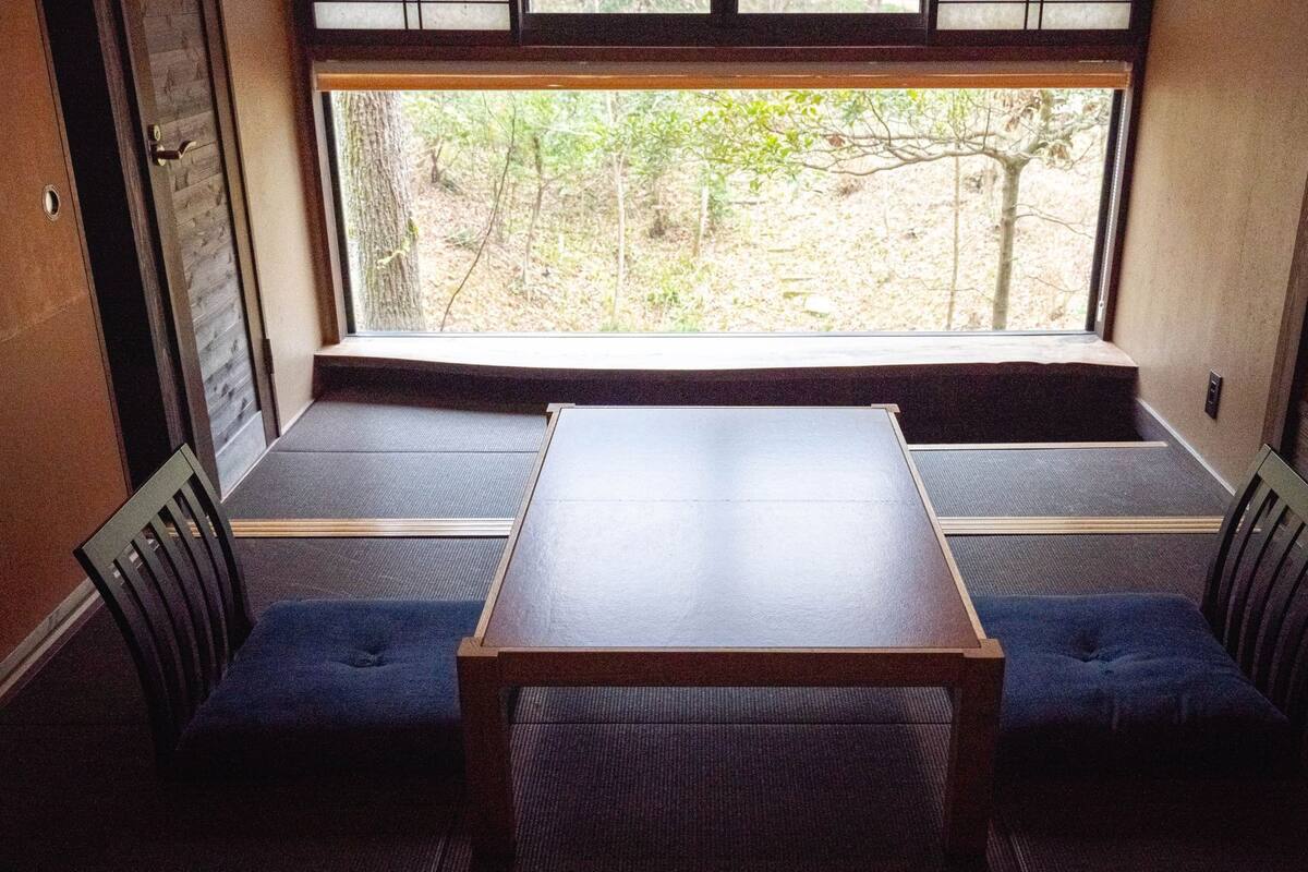 A room with a wooden table and two blue cushioned seats, a window with a view of trees, and a door with a handle.