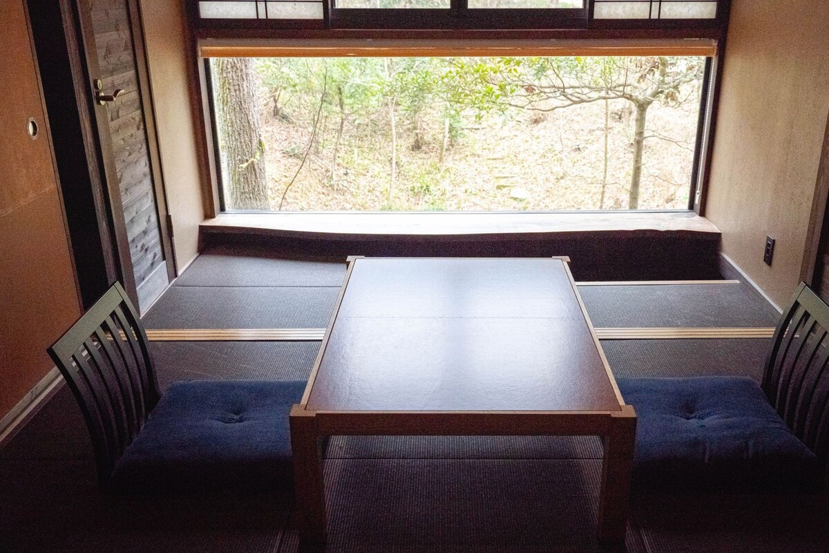 Une pièce avec une table en bois et deux sièges bleus rembourrés, une fenêtre donnant sur des arbres et une porte avec une poignée.